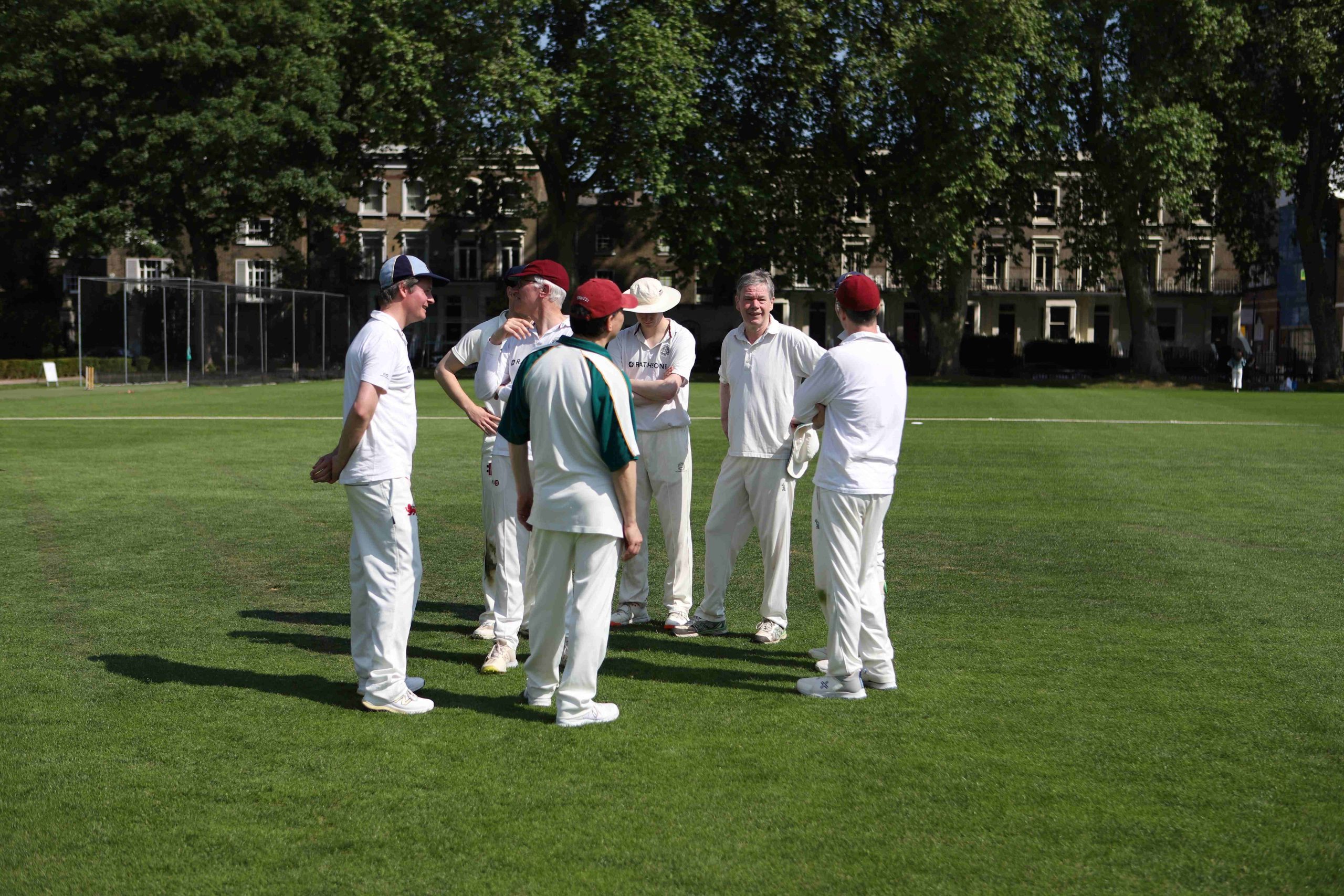 The Authors XI are standing in an informal circle chatting with various different coloured caps on, two are smiling and behind them in the distance are trees and redbrick buildings