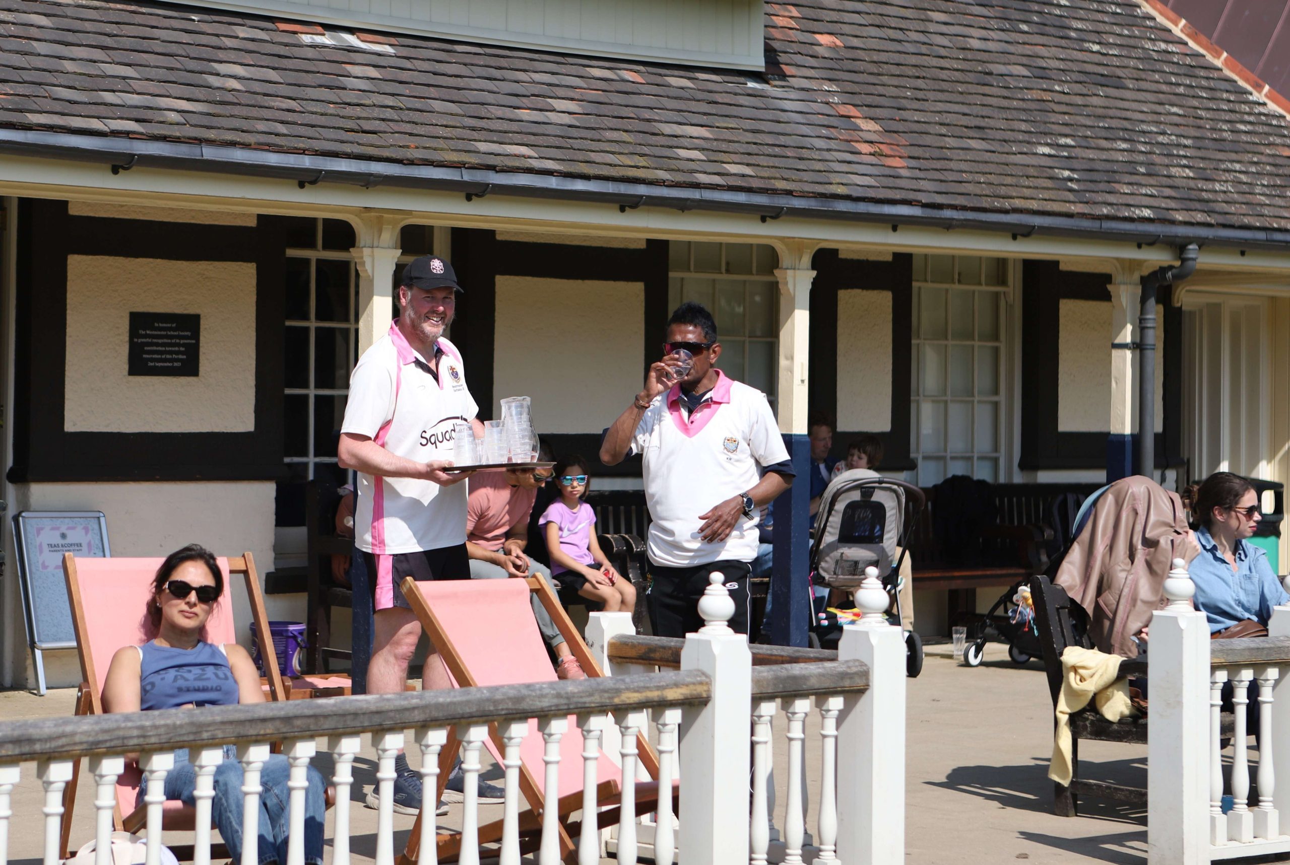A cricketeer in Westminster sports kit, pink, black and white is smiling for the camera by the pavilion after pouring another cricketer water - he is drinking and smiling at the camera too and family and friends are sat dotted around