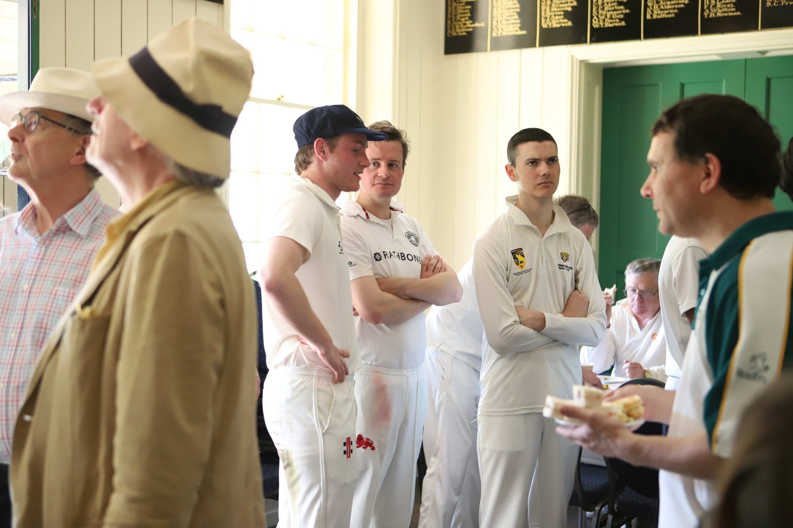 Three authors in whites inside the pavilion - two are chatting and the other is looking up at something - above them are names engraved in gold on a black wooden board and there is a window to the left