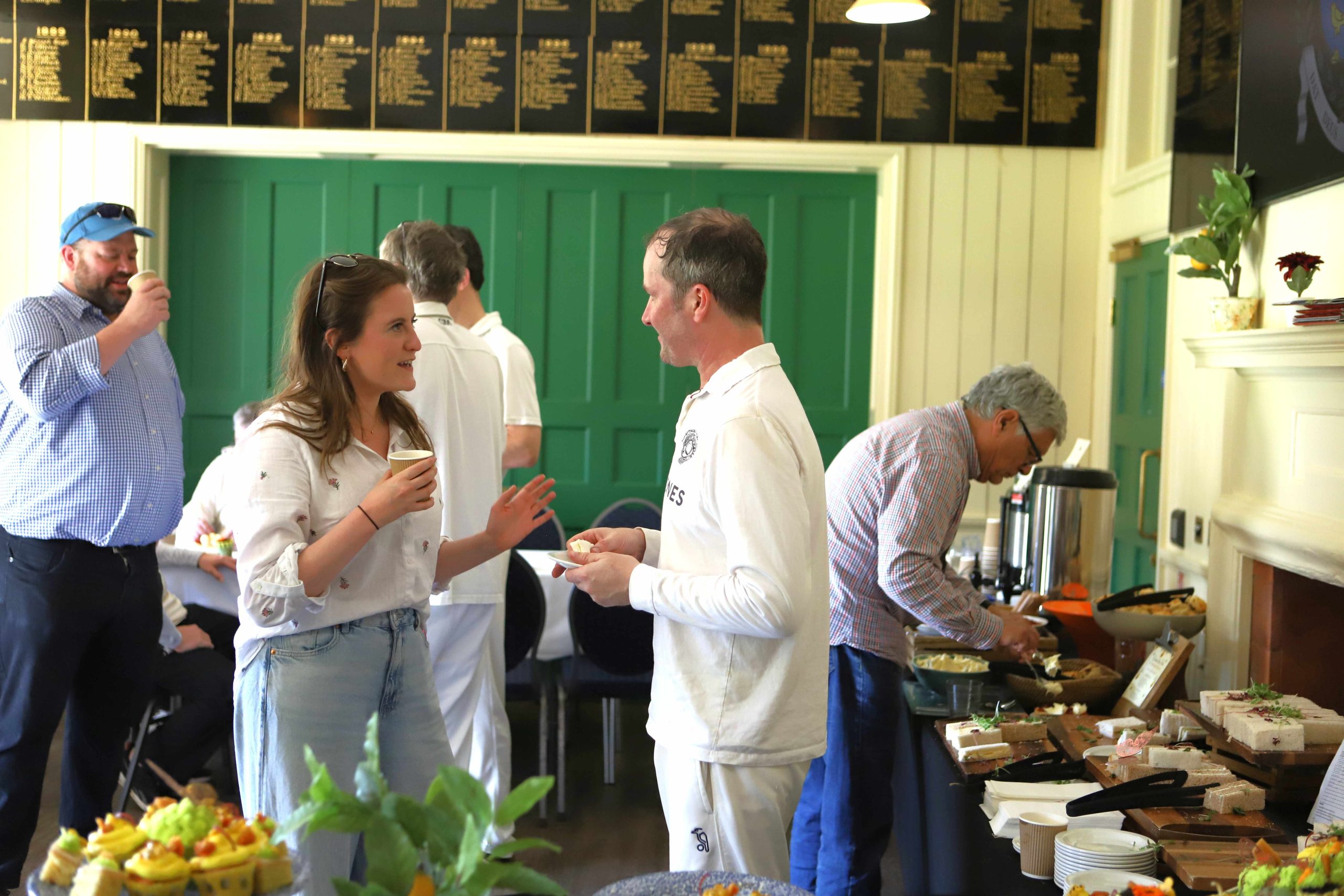 A lady in casual wear talking to an author in whites in front of the tea table, there is a man pouring tea to the right and behind are more cricketers talking near some aqua green internal wooden doors above which are lists on name engraved in gold on black painted wooden boards