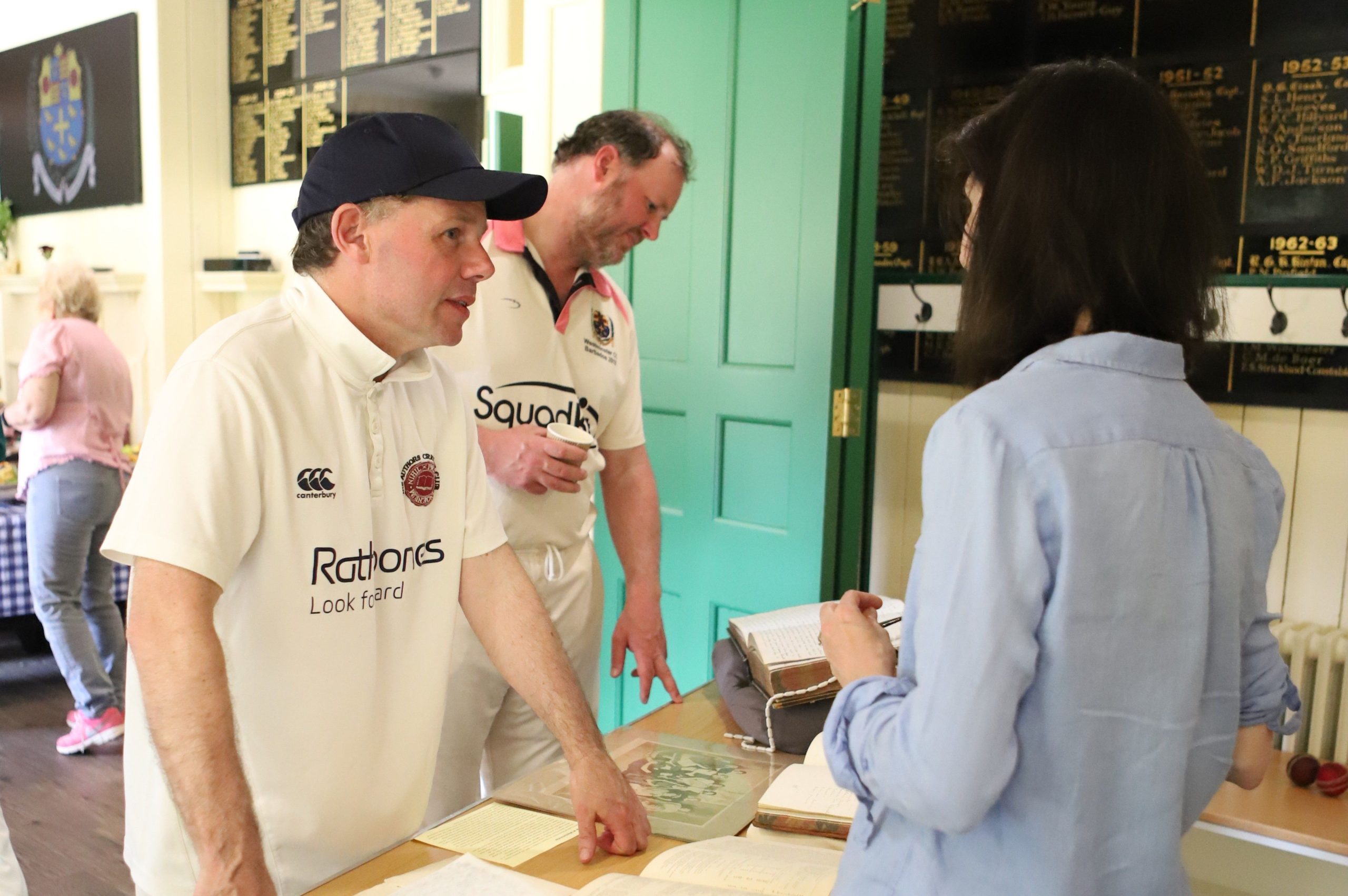 Two men in cricket whites, one in a cap nearer the camera talking to the female archivist who is wearing a light blue linen shirt, there are lots of publications open on the table between them and there is an aqua green internal wooden door open behind them