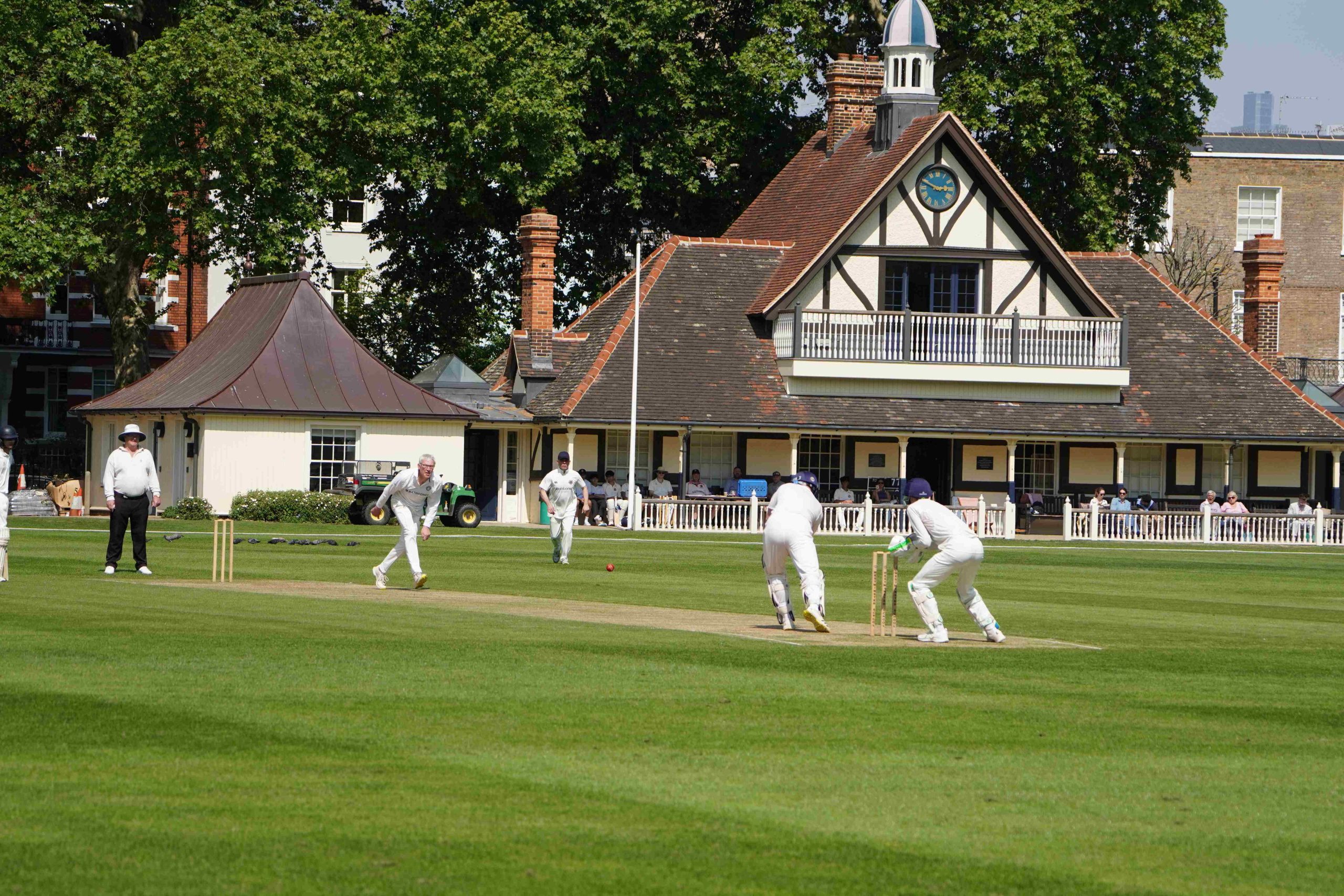 The Authors' bowler has just bowled and players are moving, the pavilion is in the background with onlookers and behind it tall green trees - the grass is bright green