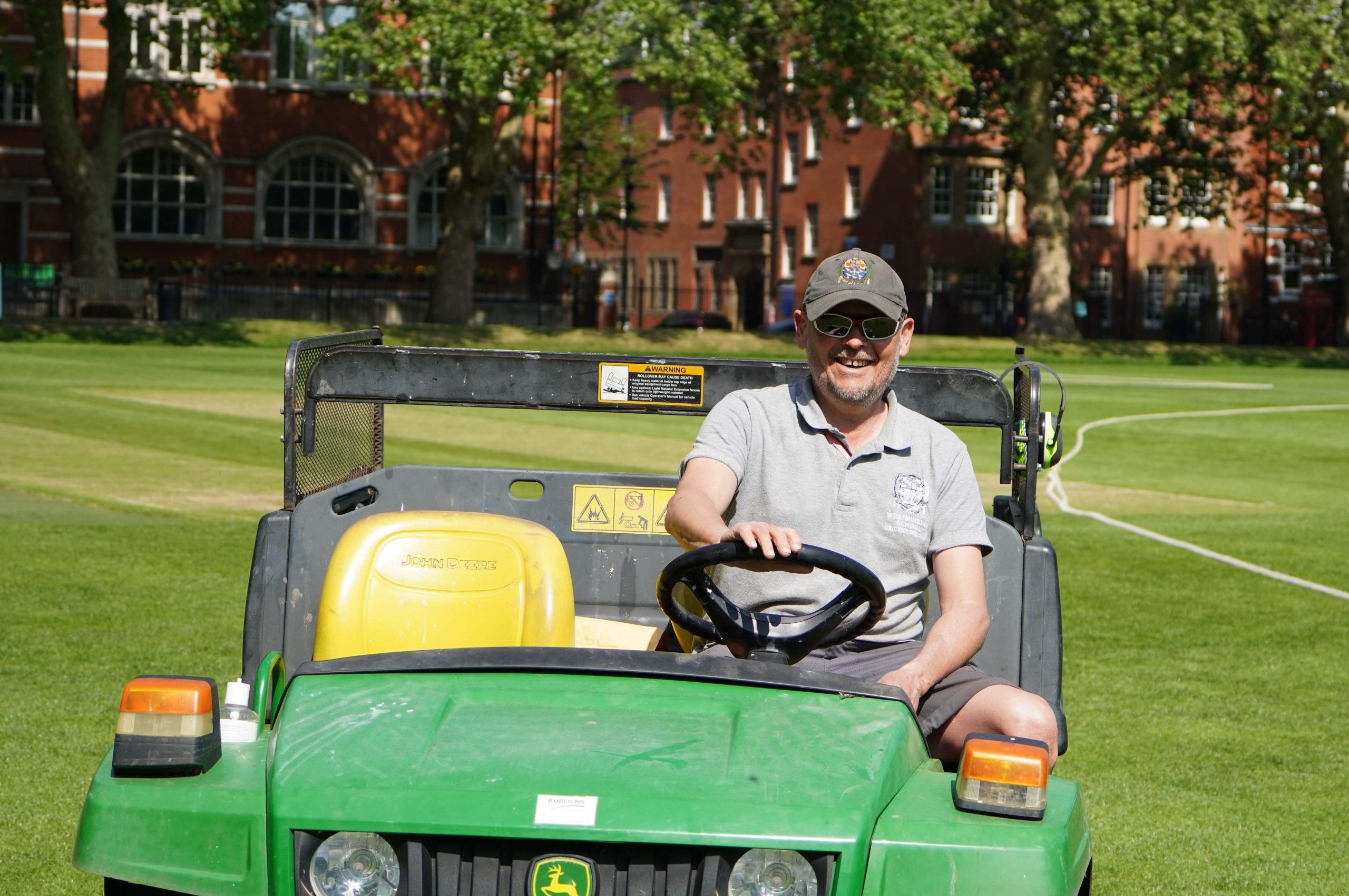 The Graoundskeeper in a grey t-shirt and Westminster cap driving his green open-top vehicle towards the camera smiling - the green grass can be seen and behind him are redbrick buildings and trees