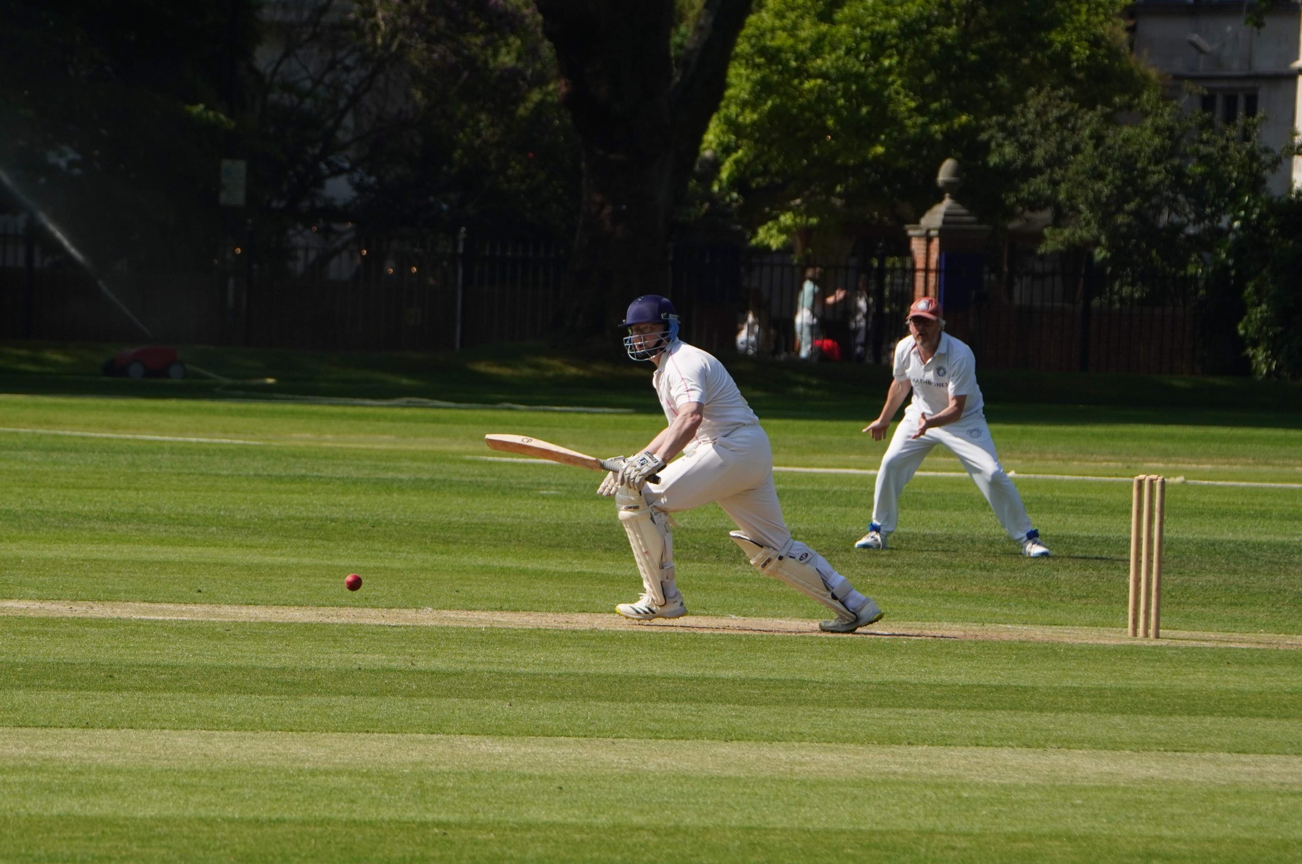 A batsman has just hit the ball and is running, the ball is to the left where he is running and a fielder in a red cap is ready to catch it