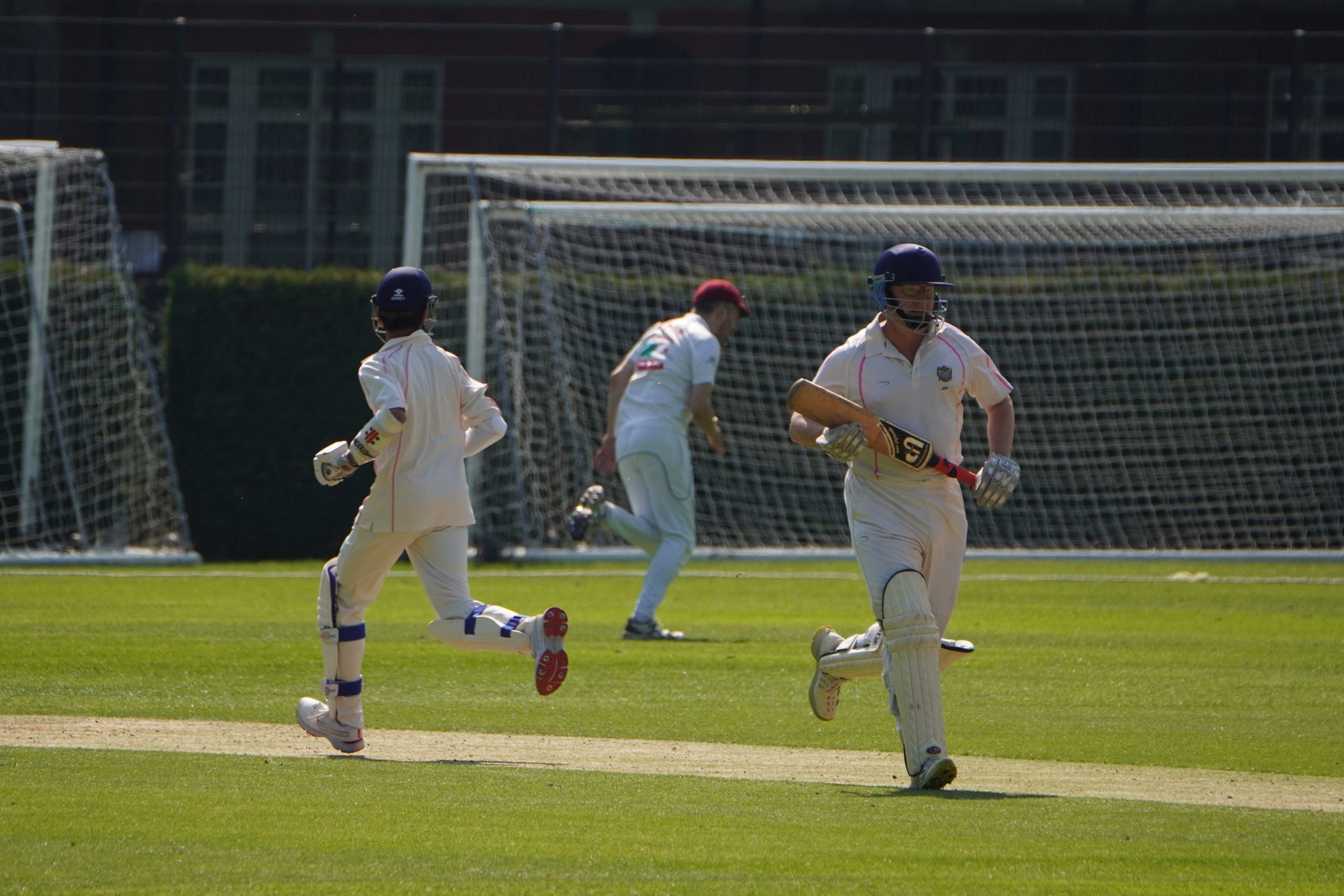 Two batters running in opposite directions a fielder in the distance between them running for the ball - there is a football goal behind him