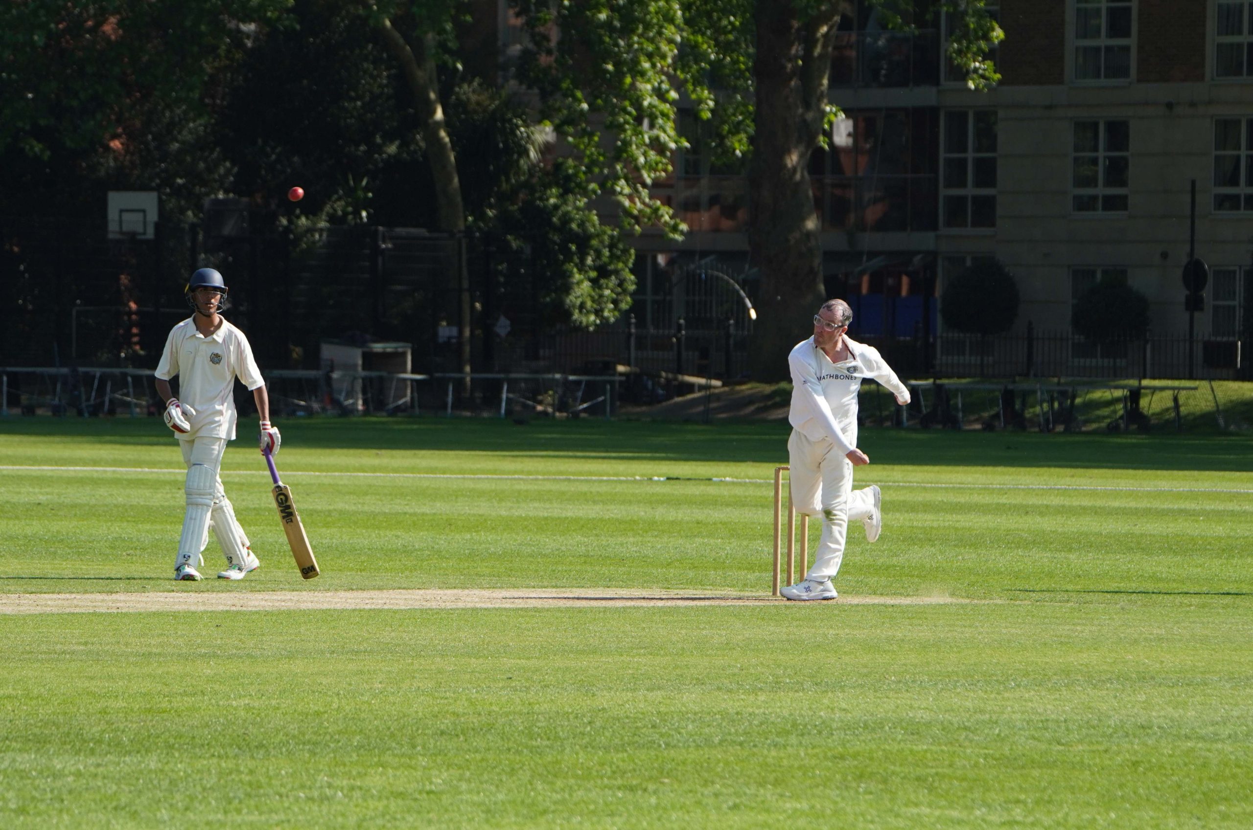 A bowler for The Authors has just bowled and has one leg up wit his arm in front, the ball is flying though the air and there are tree lined up in the distance