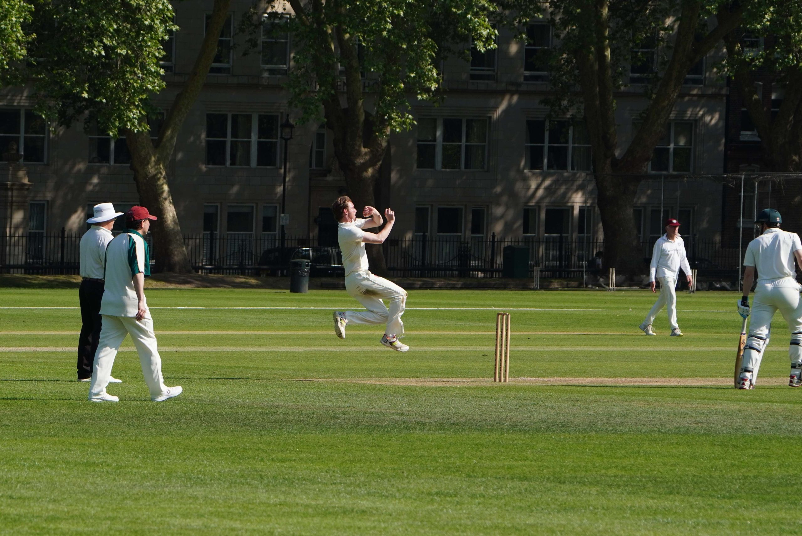 An Author in the air bowling, he has jumped and is about to swing by the wicket - there are trees in the distance