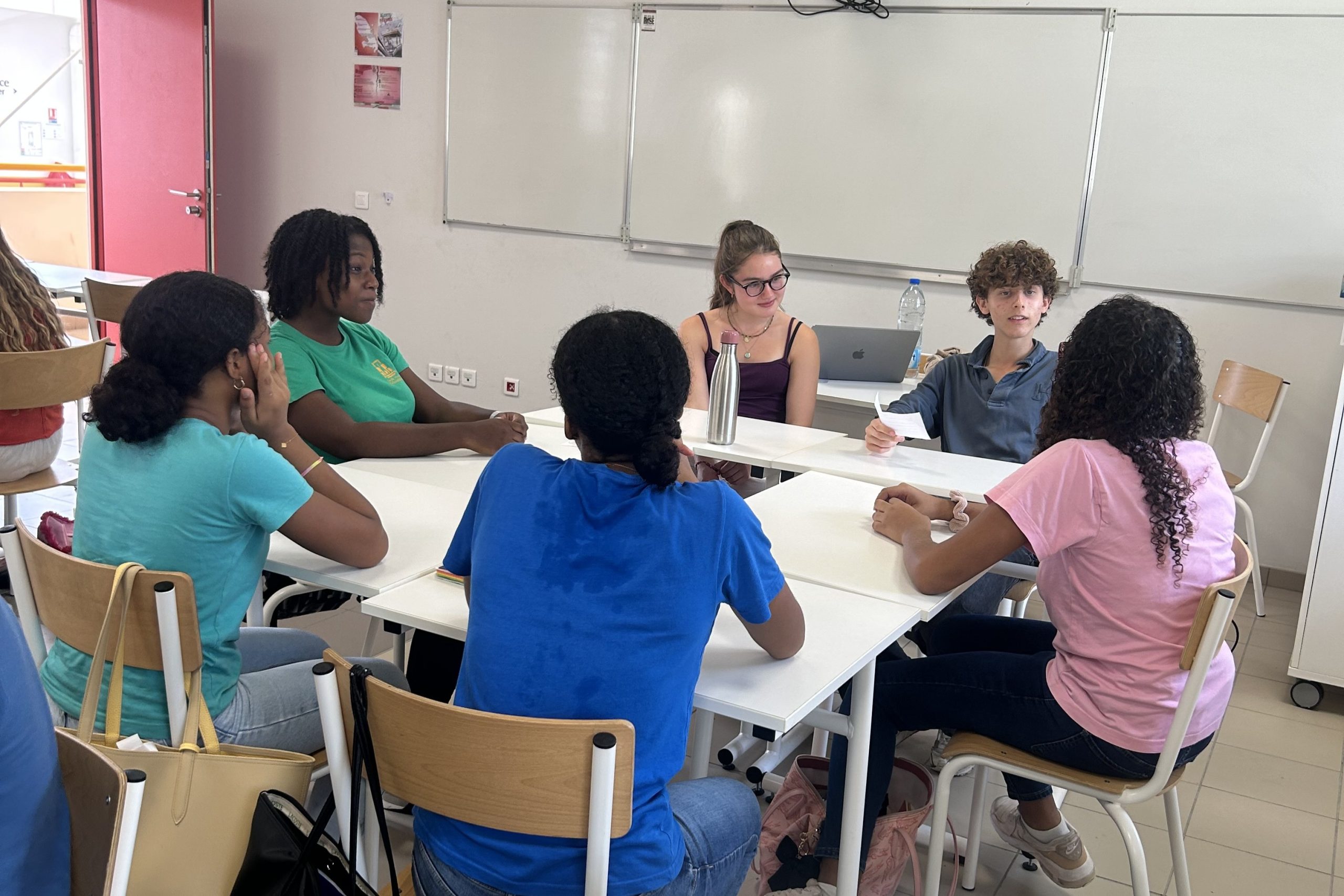 Pupil from Westminster joining pupils from Martinique at their table in a classroom - everyone is in summer clothing