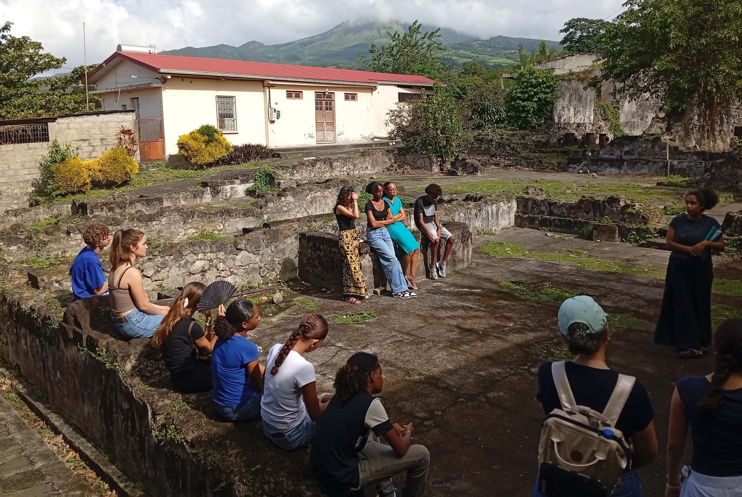 Westminster pupils sat on a wall in what looks like the foundations of the old school in Martinique, with ruble and stone and a white bungalow and mountains in the background - there are local pupils sat on the perpendicular wall