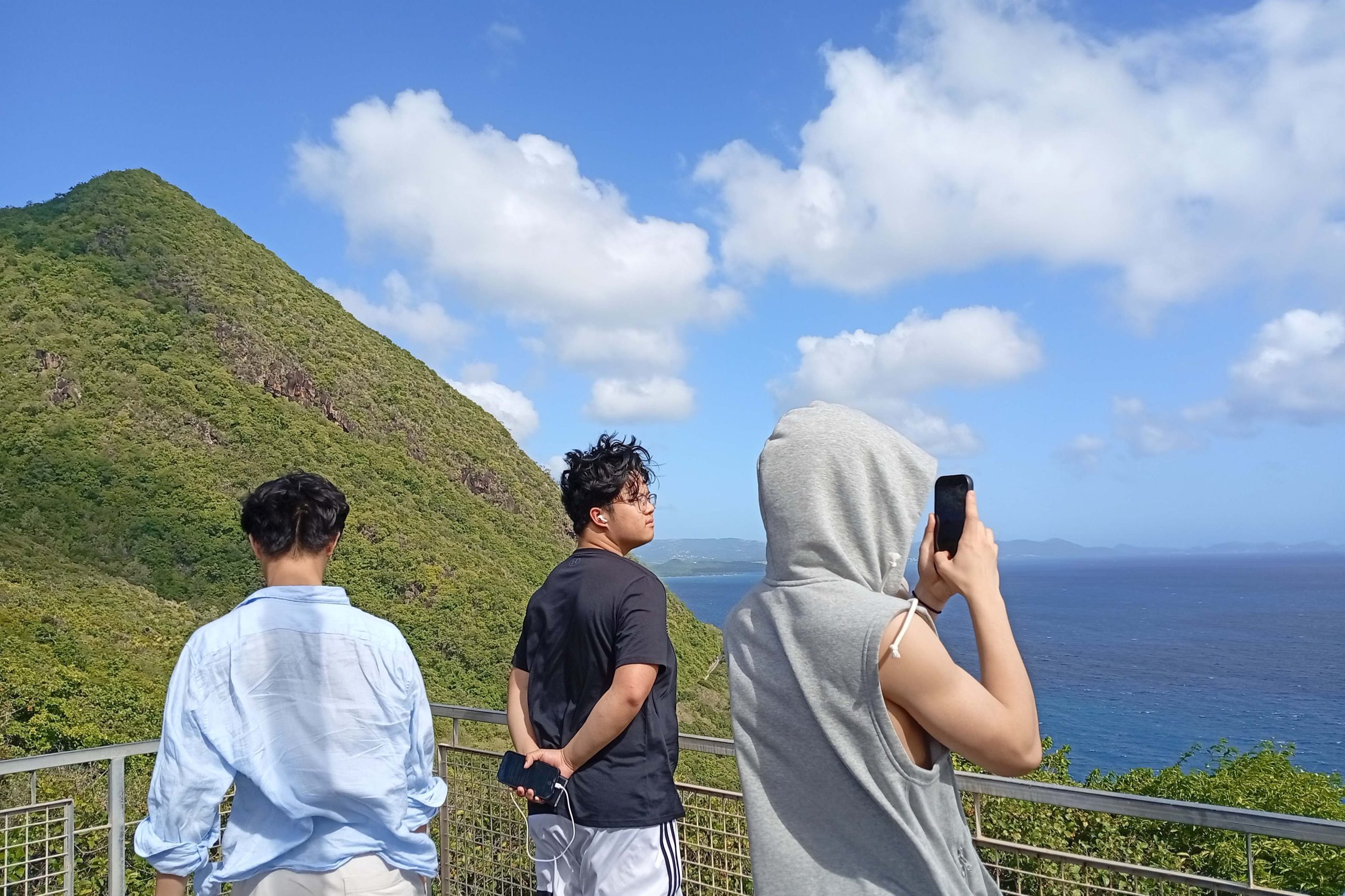Boys looking out over a very blue see with a green and lush mountain in the background, one is in a grey hoodie taking photos on his phone