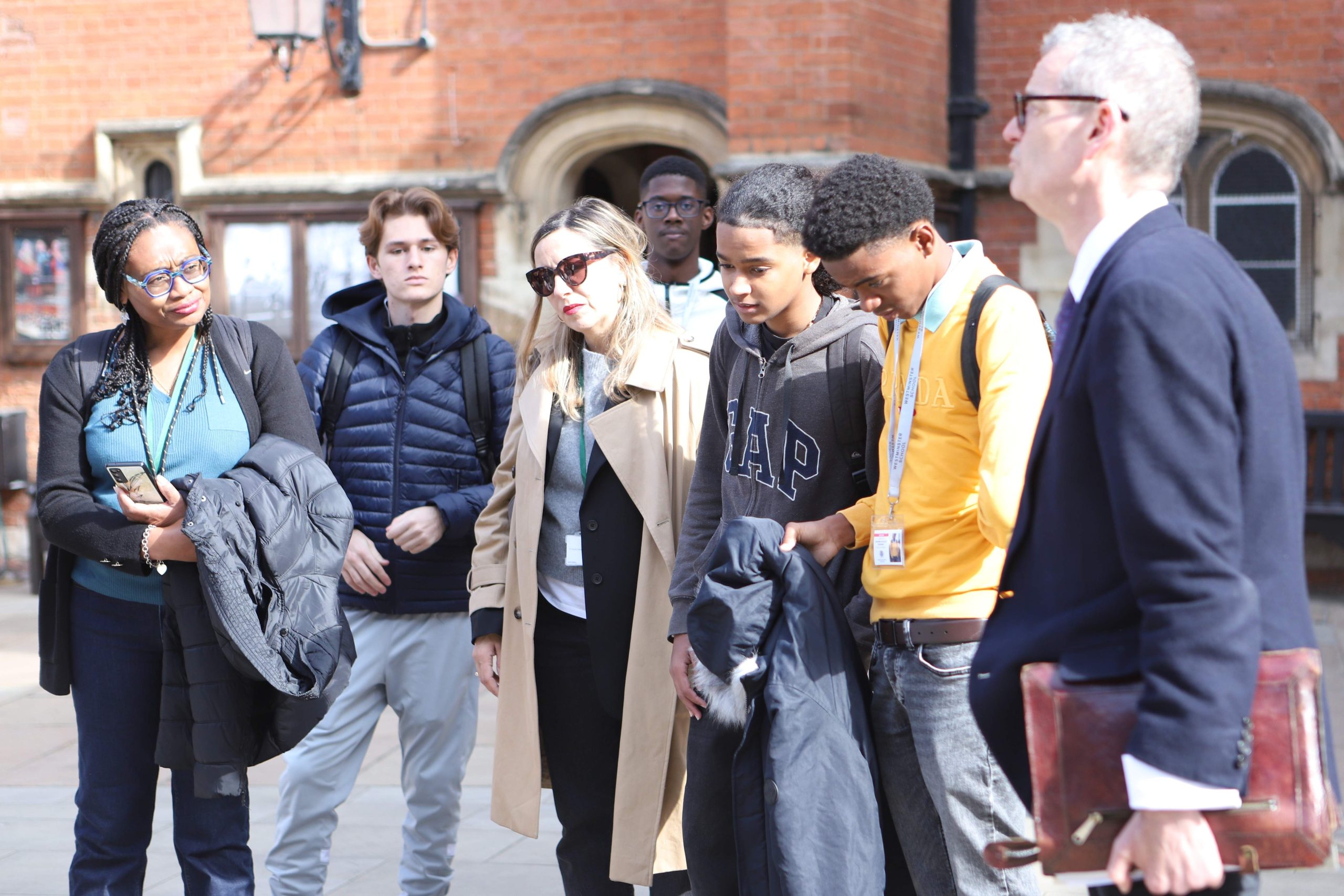 A group of pupils and teachers from Martinique looking down at the floor in Little Dean's Yard, there is a redbrick wall in the background