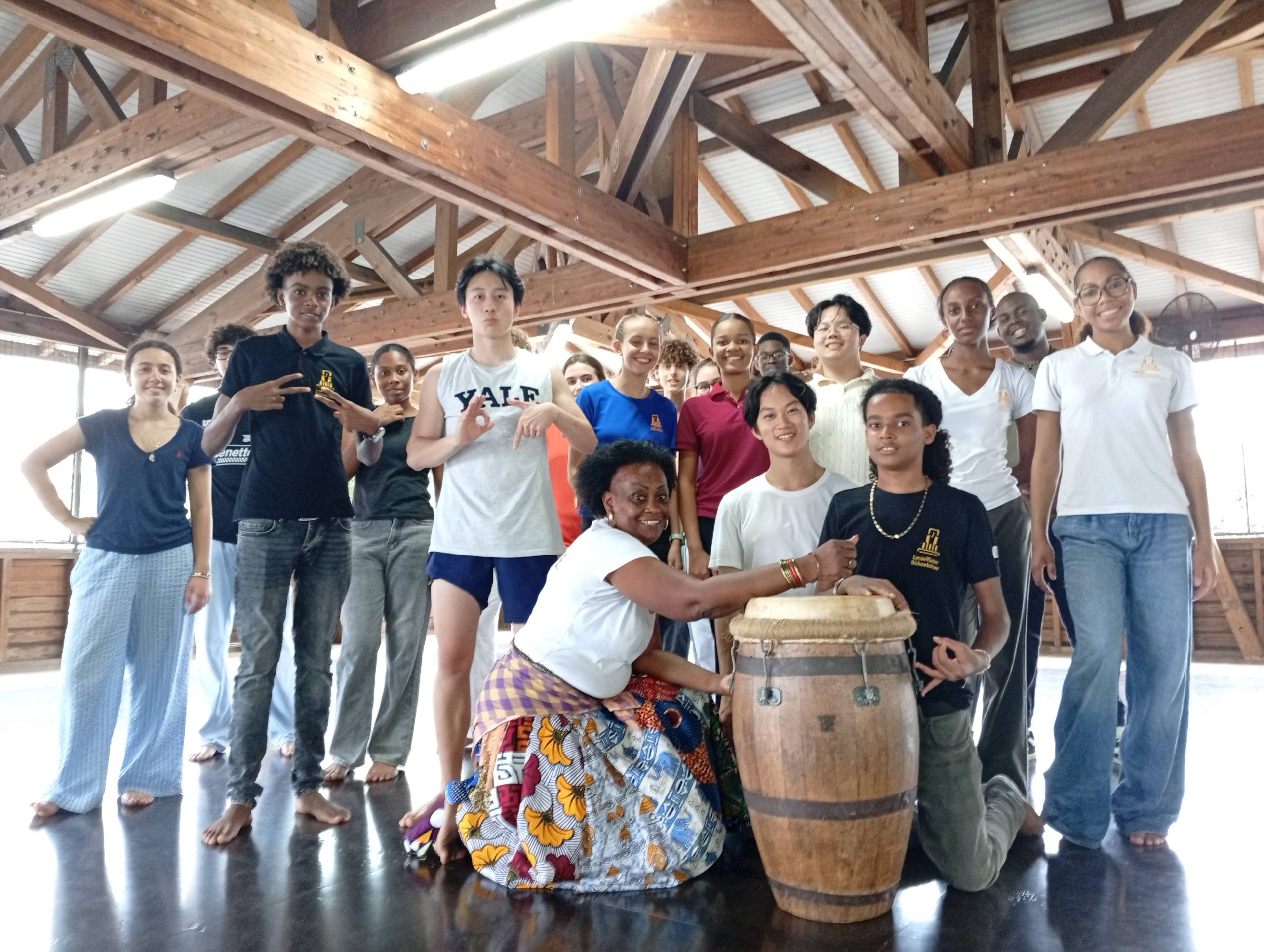 Group of Martinique and Westminster pupils standing together with a few sitting in a wooden beamed hall with a teacher and drum at the forefront smiling