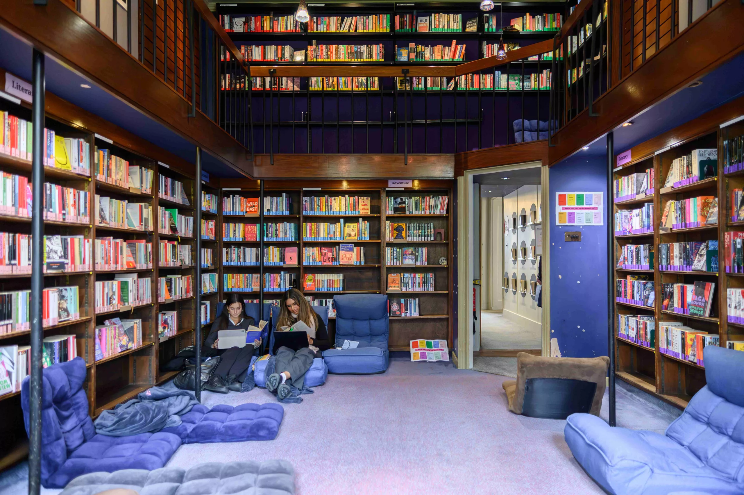 Two girls sat reading on their laptops holding books in the corner of a library space with ceiling high dark-wood bookshelves and a mezzanine level above them with more books, they are slumped on floor cushions and there is a doorway arch to the right - the carpet is lilac