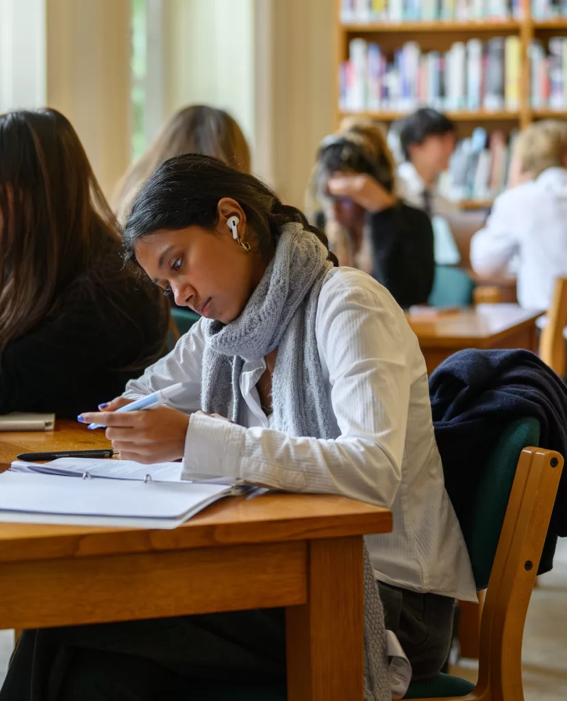 A girl in a white shirt and scarf sat writing at a shared library desk, there is a girl next to her and a table of boys and girls in the background set against a ceiling-high bookcase